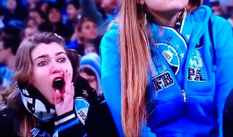 Duas jovens com uniforme do Grêmio, uma em primeiro plano à esquerda com a mão na boca e gritando e a outra, de azul, aparecendo apenas até metade do rosto