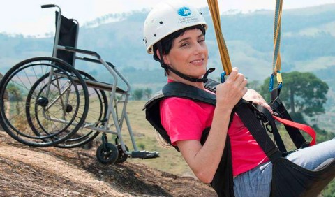 Moça de capacete em um suporte de rapel e, no plano de fundo, uma cadeira de rodas