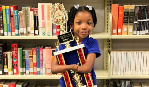 Foto horizontal de uma garotinha negra ao centro, segurando um troféu entre os braços, sorrindo. Ela não possui as mãos e, atrás dela, uma estante com vários livros.