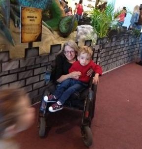 Foto vertical de uma mulher em uma cadeira de rodas com seu filho no colo. Ambos estão em um shopping, na frente de uma estátua de um dragão. Ao fundo, algumas pessoas observam a exposição