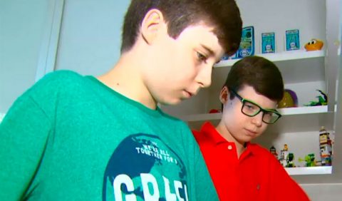 Dois irmãos gêmeos, de 10 anos, autistas, lado a lado. Em primeiro plano, Giuliano, de camisa verde; em segundo plano, Luigi, de camisa vermelha e óculos. Ao fundo, uma estante com cartões e brinquedos.