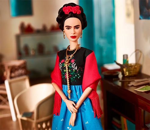 Boneca Barbie vestida de Frida Khalo, com uma saia azul, blusa preta com estampa de flores, um cordão longo e dourado no pescoço, uma flor vermelha nos cabelos e brincos dourados. Ao fundo, uma mesa com pincéis e uma cesta, uma cadeira branca e uma estante