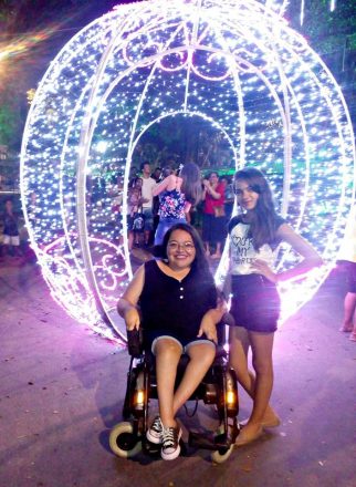 Foto vertical de duas mulheres em frente a um globo formado por muitas luzes. A mulher da esquerda está em uma cadeira de rodas e, à direita, uma criança de 12 anos está de pé, encostada na cadeira.