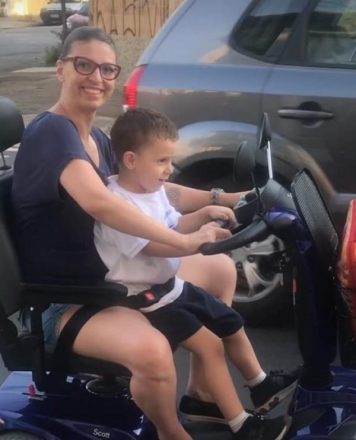 Foto vertical de uma mulher, sorrindo, de óculos, pilotando uma scooter com seu filhinho de quatro anos sentado na sua frente.